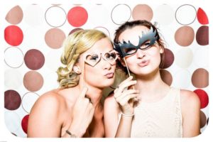 two women hold up masks to their face against a photo booth background.